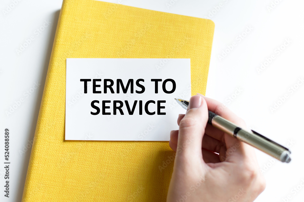 Terms to Service text is written by a person's hand on a card that lies on a yellow notebook on a light desk Terms to Service text is written by a person's hand on a card that lies on a yellow notebook on a light desk