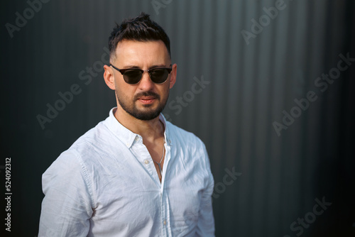 Fashion model handsome young man with trendy hairstyle in sunglasses in white shirt in light jeans posing near vintage wall on street on sunny day. Attractive american guy on walk. Copy space.