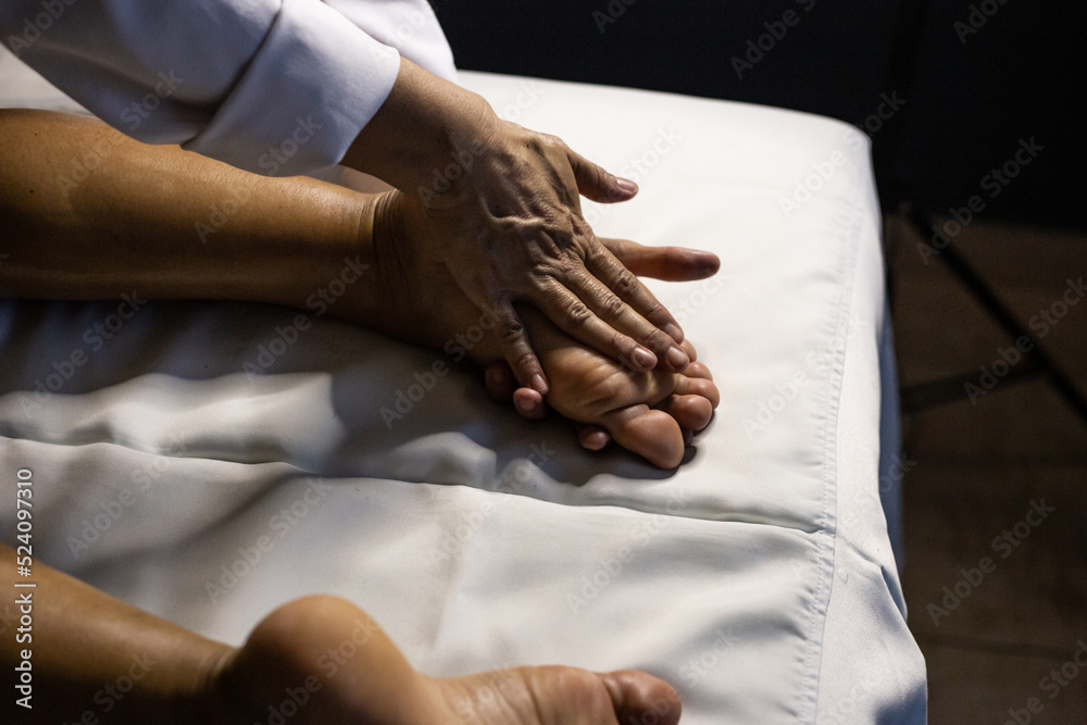 Detalhe das mãos de massagista aplicando massagem terapêutica no pé de um paciente que está deitado em uma maca com lençol branco.