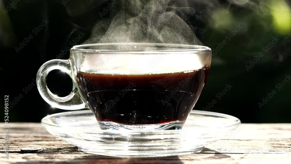 Close up hot coffee in clear glass coffee cup, mug on old wooden table in morning sunlight nature background. Hot coffee or tea cup with beautiful steaming smoke, slow motion. Hot Drink Concept Close up hot coffee in clear glass coffee cup, mug on old wooden table in morning sunlight nature background. Hot coffee or tea cup with beautiful steaming smoke, slow motion. Hot Drink Concept