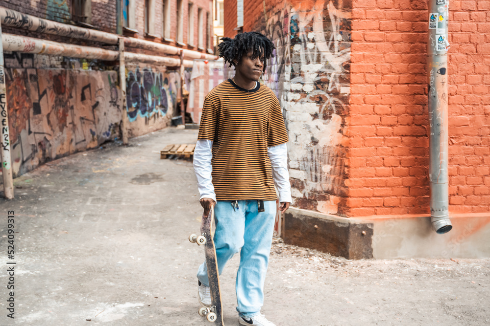 Young handsome stylish black man with natural hair dreadlocks. Afroamerican guy.Stairs,wall painted with graffiti in poor quarter of street art culture city district.African american skateboarder man