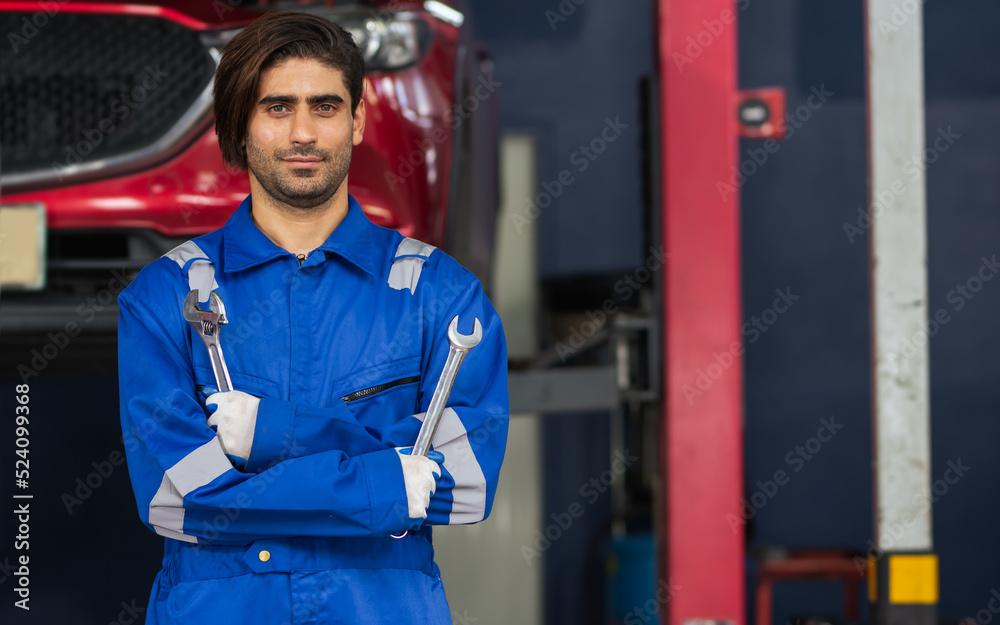 Fototapeta premium Adult handsome male mechanic wearing uniform, crossed arms, posing with confidence, standing in garage at car or automobile maintenance service center or shop with copy space. Industry Concept