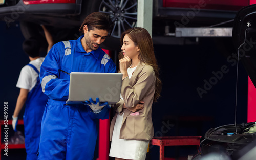 Handsome male mechanic wearing uniform, using laptop, checking or inspecting for fix, repair car or automobile components, talking with customer, working in car maintenance service center or shop.