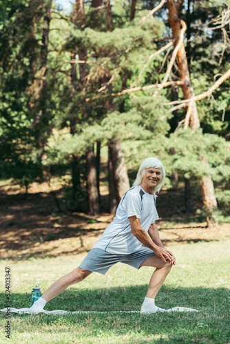 full length of senior man with grey hair smiling and doing lunges on fitness mat in park.
