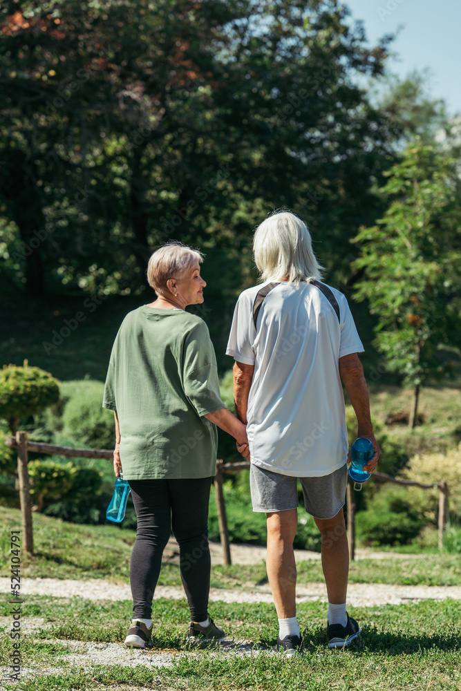 happy retired woman in sportswear looking at husband with sport bottle and holding hands in park. happy retired woman in sportswear looking at husband with sport bottle and holding hands in park.