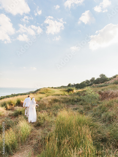  The groom in a beige suit and the bride in a white dress with a long veil are walking along a hill overgrown with steppe grasses. The sea is in the background. 