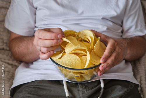 A man eats crispy potato chips from a transparent bowl on the couch. Quick snack. Calories and diet