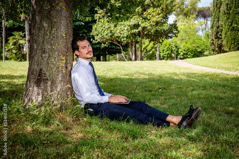 The beautiful business man is working in a park.The young business man is sitting on the lawn and leaning against a tree while he is writing a work email. The beautiful business man is working in a park.The young business man is sitting on the lawn and leaning against a tree while he is writing a work email.