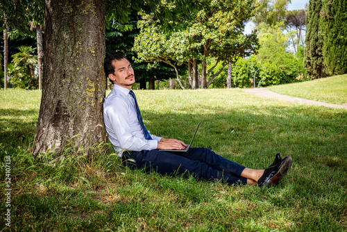 The beautiful business man is working in a park.The young business man is sitting on the lawn and leaning against a tree while he is writing a work email.
