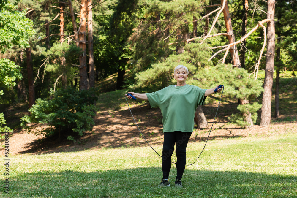 full length of senior woman with grey hair smiling and exercising with jumping rope in park.
