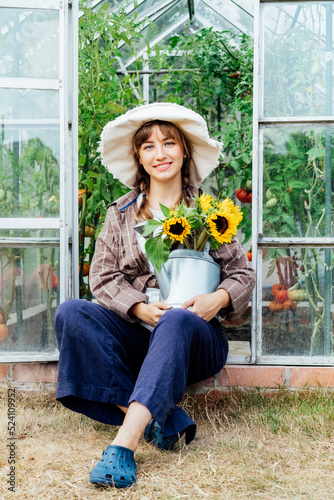 Portrait of smiling young female farmer woman holding watering can with fresh sunflowers bouquet, sitting near greenhouse. Cottagecore lifestyle. Growing organic vegetables in garden. Vertical card photo