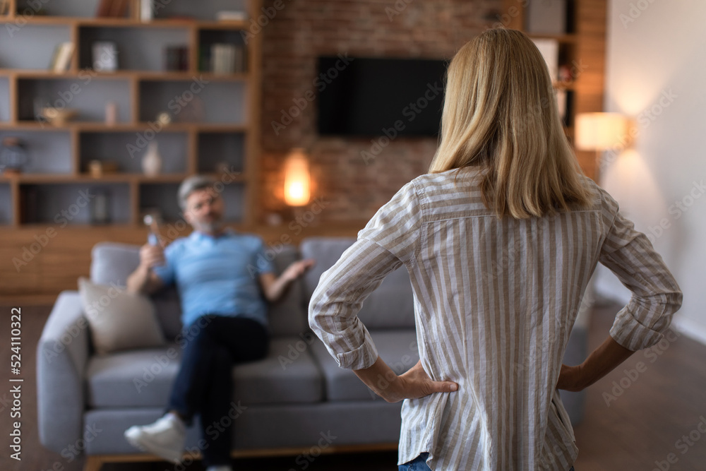 Unhappy sad adult caucasian wife angry at drunk husband with bottle in living room interior, back, blurred