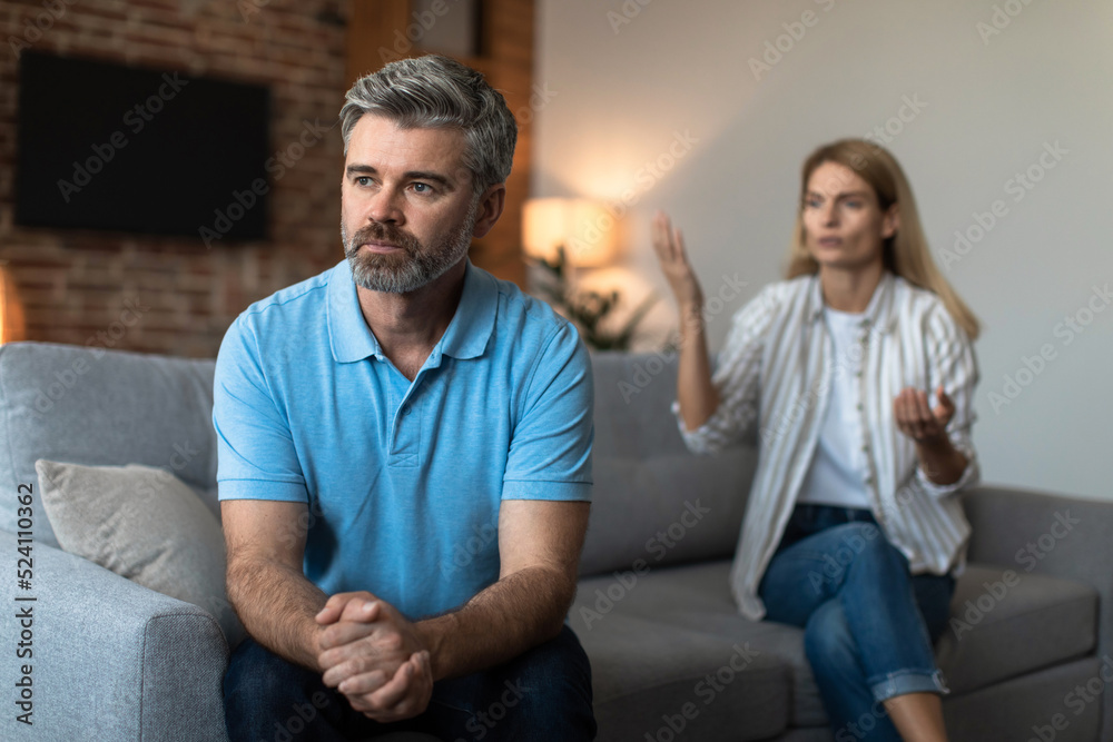 Offended unhappy mature caucasian woman scolds at sad husband, man ignores wife in living room interior Offended unhappy mature caucasian woman scolds at sad husband, man ignores wife in living room interior