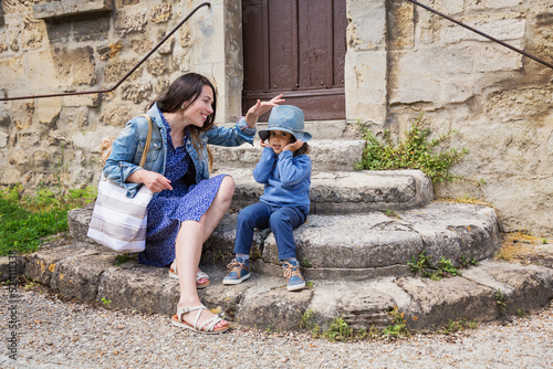 Mother and little handsome baby boy sitting on ancient stone stairs and playing outdoor with straw hat in old town