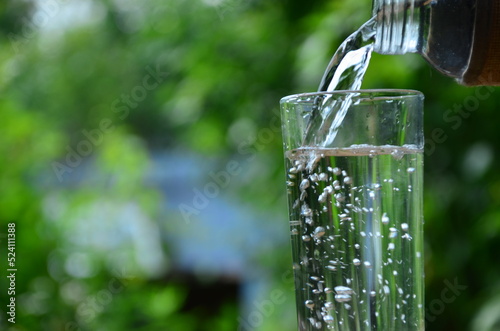 Drink water pouring in to glass outdoor over sunlight and natural green background on white table.