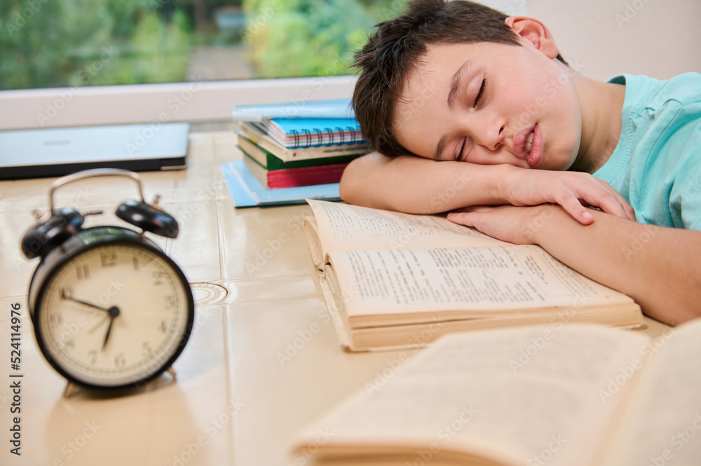 Blurred black alarm clock on the background of a tired sleepy schoolboy falling asleep on a textbook while doing homework. Teenage boy having learning disability. Back to school Blurred black alarm clock on the background of a tired sleepy schoolboy falling asleep on a textbook while doing homework. Teenage boy having learning disability. Back to school