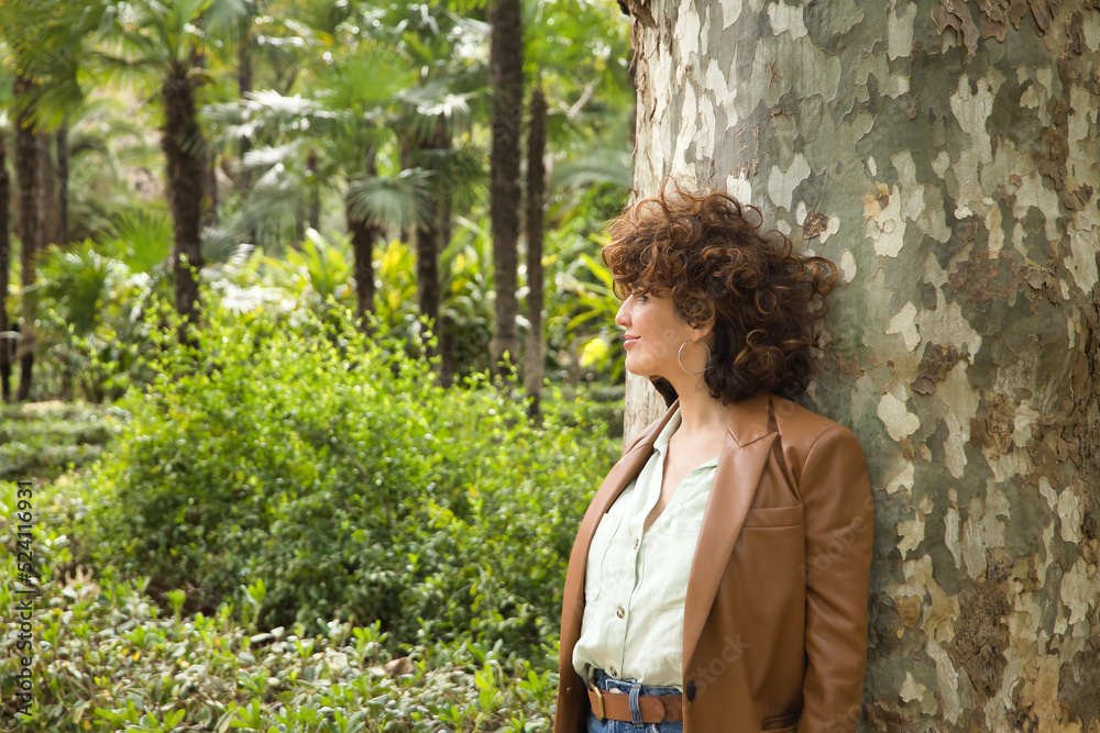 Attractive mature woman with curly brown hair, wearing a brown leather jacket and jeans leaning against a tree trunk, pensive and dreamy. Concept thought, dreams, hairstyle, curls, nature. Attractive mature woman with curly brown hair, wearing a brown leather jacket and jeans leaning against a tree trunk, pensive and dreamy. Concept thought, dreams, hairstyle, curls, nature.