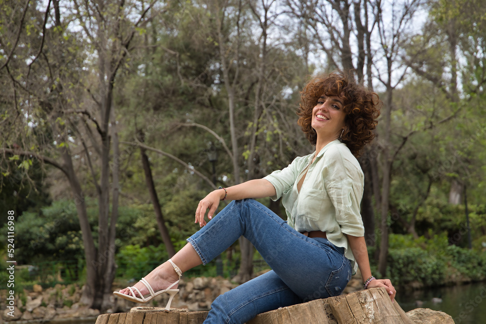 Portrait of a mature and attractive woman, with curly brown hair and light green shirt, sitting on the cut trunk of a tree by a lake. Concept beauty, fashion, trend, lake, hairstyle, curls. Portrait of a mature and attractive woman, with curly brown hair and light green shirt, sitting on the cut trunk of a tree by a lake. Concept beauty, fashion, trend, lake, hairstyle, curls.