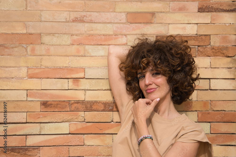 Portrait of a mature and attractive woman, with curly brown hair and brown T-shirt on a brick background, with her hand on her chin, thoughtful. Concept of thinking, meditating, doubting, combing.