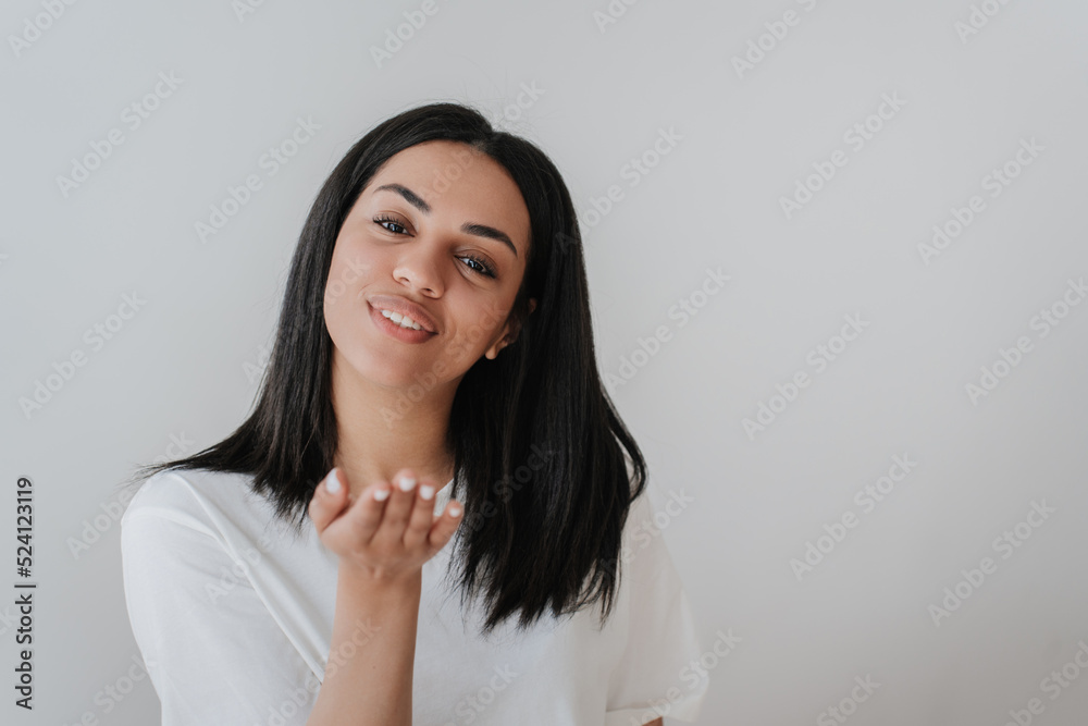 Adorable African American woman in white t-shirt sending air kiss, looking at camera, smiling on grey background with empty space. Satisfied Brazilian girl isolated. Mockup beautiful people in love. Adorable African American woman in white t-shirt sending air kiss, looking at camera, smiling on grey background with empty space. Satisfied Brazilian girl isolated. Mockup beautiful people in love.