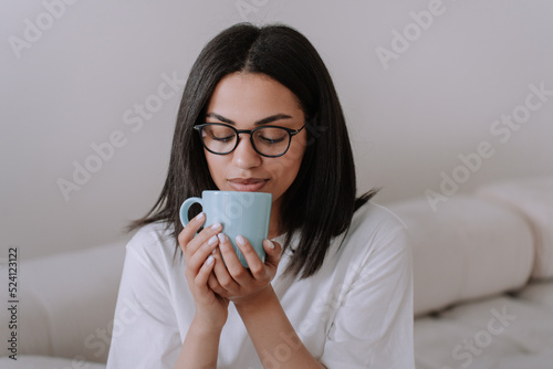 Adorable african american female in glasses and white t-shirt holding cup of coffee, sitting home on sofa looking at camera, toothy smiling.Brazilian young woman at home. Mockup beautiful girl.