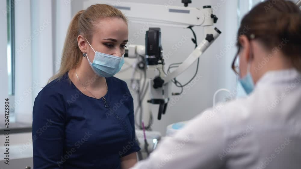 Portrait of focused dental assistant sitting in hospital with doctor examining patient in slow motion. Concentrated Caucasian young woman in face mask assisting professional doctor indoors Portrait of focused dental assistant sitting in hospital with doctor examining patient in slow motion. Concentrated Caucasian young woman in face mask assisting professional doctor indoors