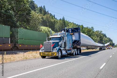 Big rig semi truck tractor with oversize load sign on the front transporting windmill electric generator blade with special additional trolley standing on the highway shoulder with escort vehicles photo