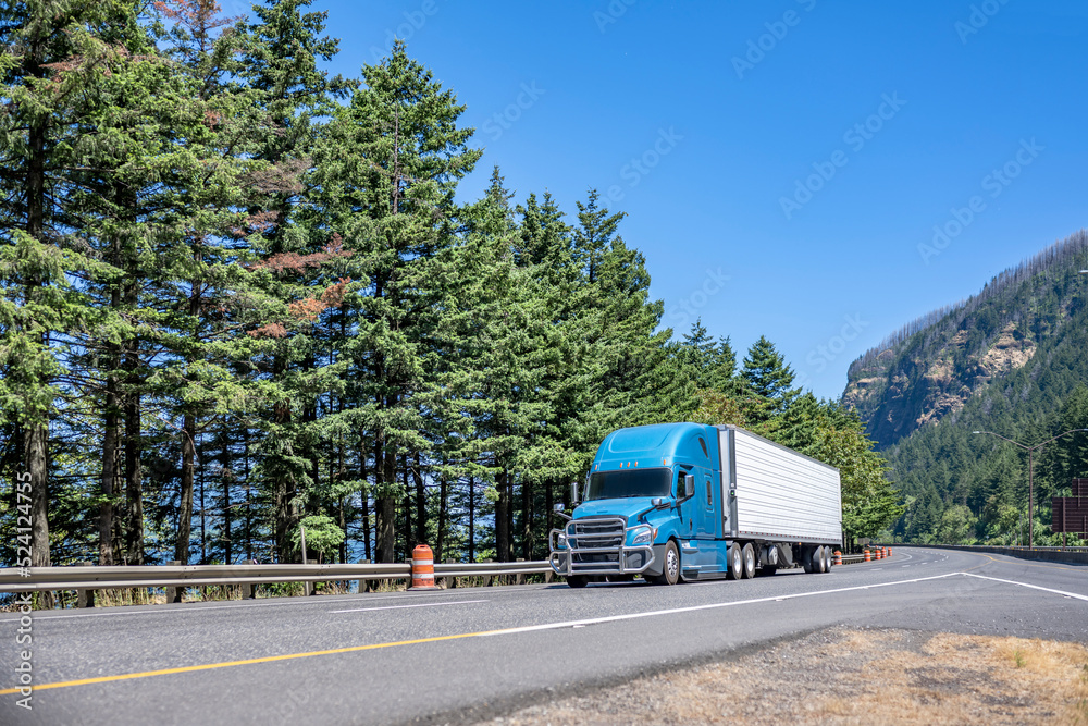 Light blue big rig semi truck with grille guard transporting cargo in refrigerator semi trailer driving on highway road with fork divided traffic lines