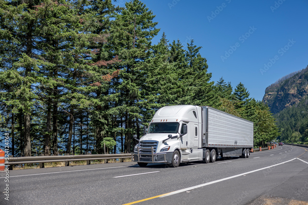 Popular pro big rig white semi truck with chrome grille guard transporting cargo in refrigerated semi trailer driving on the divided highway road Popular pro big rig white semi truck with chrome grille guard transporting cargo in refrigerated semi trailer driving on the divided highway road