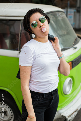 young greaser girl with short hair, posing with a fifties rockabilly style while chewing gum dressed in a white t-shirt black pants leather jacket and sunglasses with an old green hippie van (kombi) 