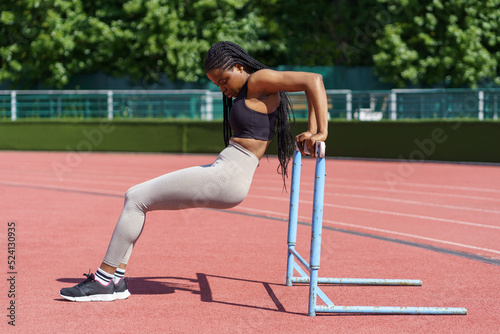 Young African American woman enjoys intensive workout on city stadium against park with lush green trees. Sportive female in sportswear pumps up legs holding on to metal barrier standing on red track