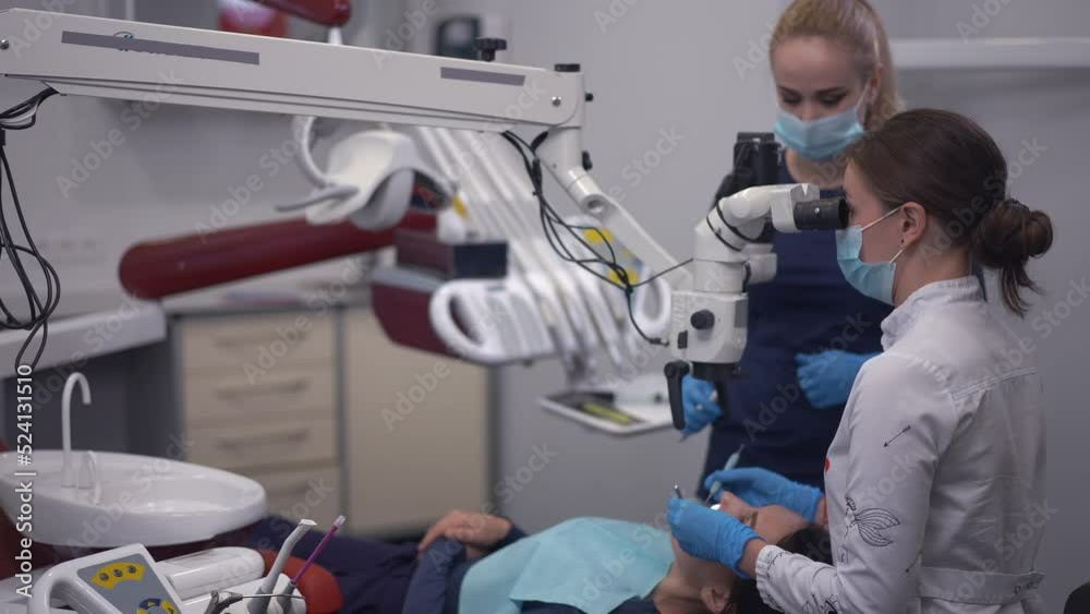 Concentrated expert dentist using microscope with assistant passing tools on the right as patient sitting in dental chair. Professional Caucasian doctor and nurse treating woman in hospital
