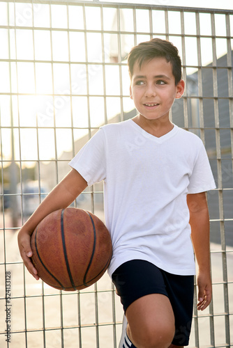 A cute young boy plays basketball on the street playground in summer. Teenager in a white t-shirt with orange basketball ball outside. Hobby, active lifestyle, sports activity for kids
