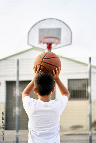 A cute young boy plays basketball on the street playground in summer. Teenager in a white t-shirt with orange basketball ball outside. Hobby, active lifestyle, sports activity for kids