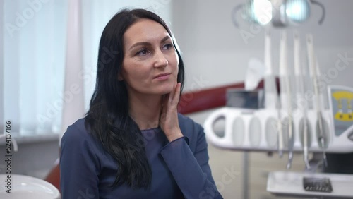 Woman touching jaw talking complaining tooth pain sitting in dental chair in hospital. Portrait of unwell Caucasian female patient consulting professional in dental clinic. Slow motion photo