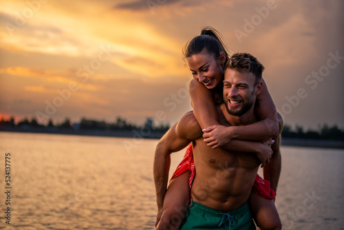 Happy young couple in love having fun relaxing on beach near city. Caucasian girlfriend piggybacking on boyfriend enjoying holidays together. Selective focus