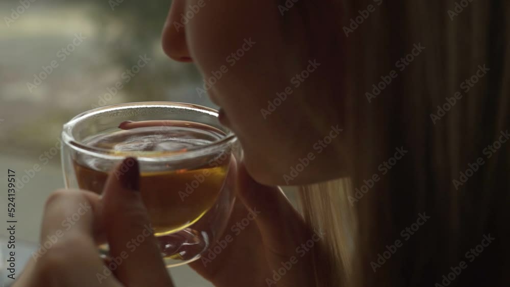A young girl is sitting near the window and holding a glass mug with warm tea blowing cool air into her cup A young girl is sitting near the window and holding a glass mug with warm tea blowing cool air into her cup