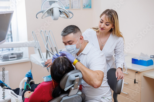 The dentist doctor looks at the patient s teeth and holds dental instruments near the mouth. The assistant helps the doctor. They wear white uniforms with masks and gloves. Dentist. Dental office