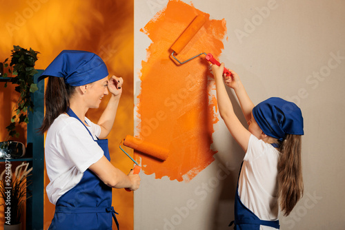 Mom and kid painting walls with orange color paint, using renovating tools and paintbrush to redecorate apartment room. Small family redecorating home interior with roller brush.