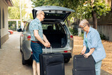 Elderly husband and wife going on holiday vacation and putting luggage or travelling bags in automobile trunk. People leaving on retirement trip journey with suitcases and trolley in vehicle.