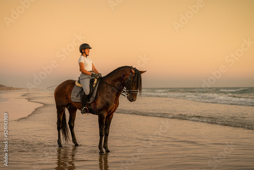 a smiling 21-year-old rider standing on the shore with her horse facing the sea
