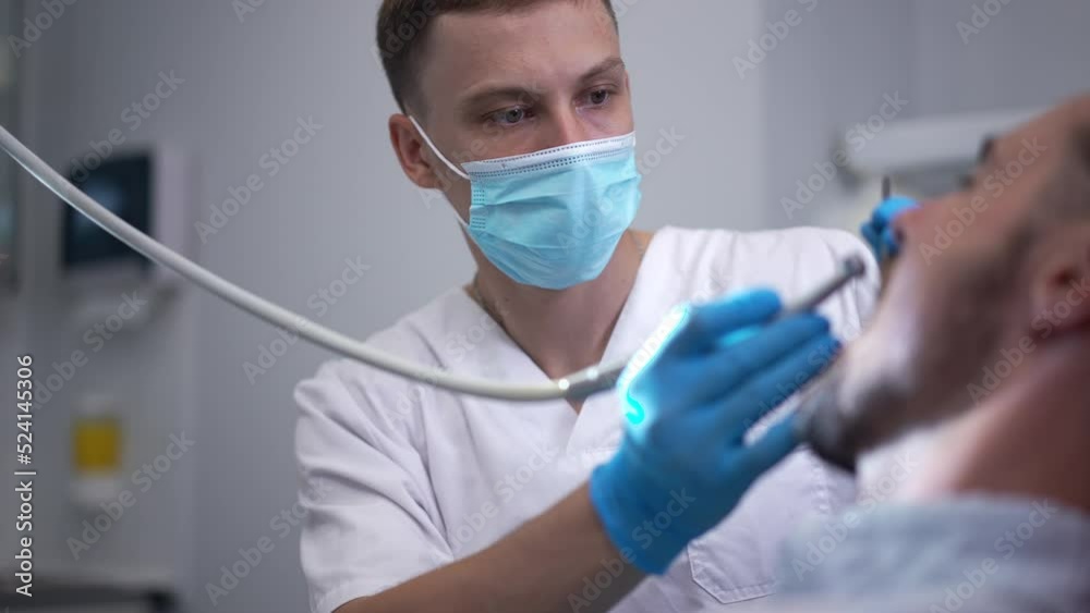 Portrait of expert dentist using dental drill with patient sitting in dental chair in hospital. Concentrated intelligent Caucasian young doctor helping man with toothache in medical clinic Portrait of expert dentist using dental drill with patient sitting in dental chair in hospital. Concentrated intelligent Caucasian young doctor helping man with toothache in medical clinic