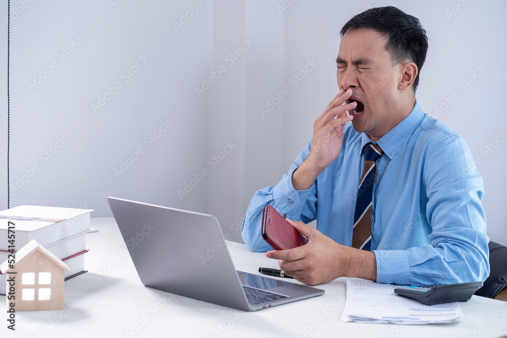 Shot of a young businessman yawning at work in office. Exhausted young businessman yawning. tired bored businessman at workplace near laptop and yawning Shot of a young businessman yawning at work in office. Exhausted young businessman yawning. tired bored businessman at workplace near laptop and yawning