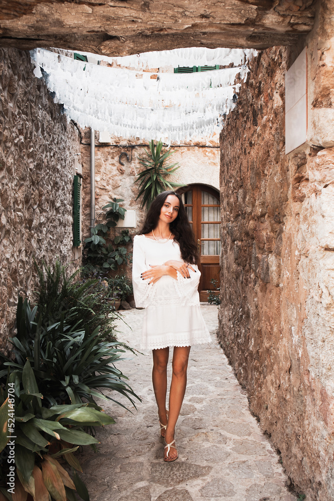 Cute charming girl, Spain, Mallorca. Cinematic Processing. Attractive brunette with curly hair woman wears white dress. Cozy street, Valldemossa, Mallorca (Majorca)