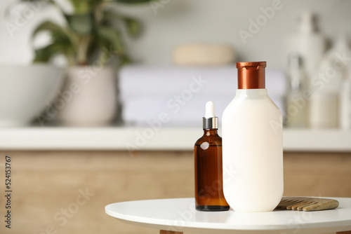 Bottle of shampoo, hair oil and wooden comb on white table in bathroom, space for text