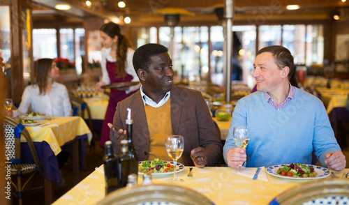 African-american and caucasian men drinking wine and talking in restaurant.