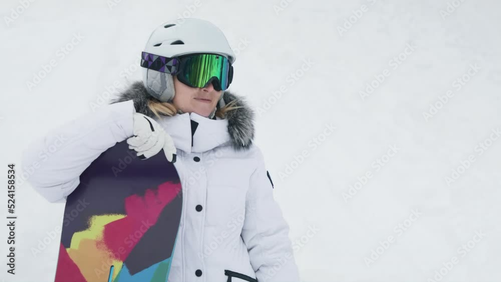 Happy young woman in stylish sportswear, white helmet, colorful goggles, snowboard with white mittens relaxing outdoors in ski resort park in winter. Slow motion smiley portrait of snowboarder girl Happy young woman in stylish sportswear, white helmet, colorful goggles, snowboard with white mittens relaxing outdoors in ski resort park in winter. Slow motion smiley portrait of snowboarder girl
