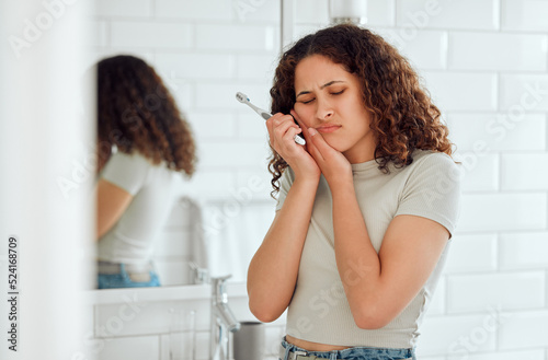 Toothache, pain and sensitive teeth with a woman brushing her teeth in a bathroom at home. Young female with a cavity suffering from discomfort during dental hygiene routine. Lady with a sore mouth photo