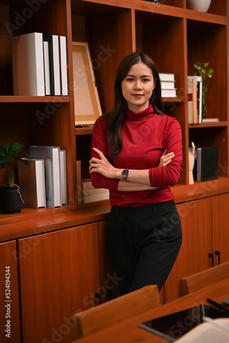 Successful Asian businesswoman stands with her arms crossed in her classic luxury private office.