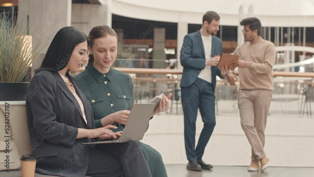Medium slowmo of two young attractive diverse female colleagues using laptop while working on project at modern business center. Two men looking at tablet pc and talking at blurred background